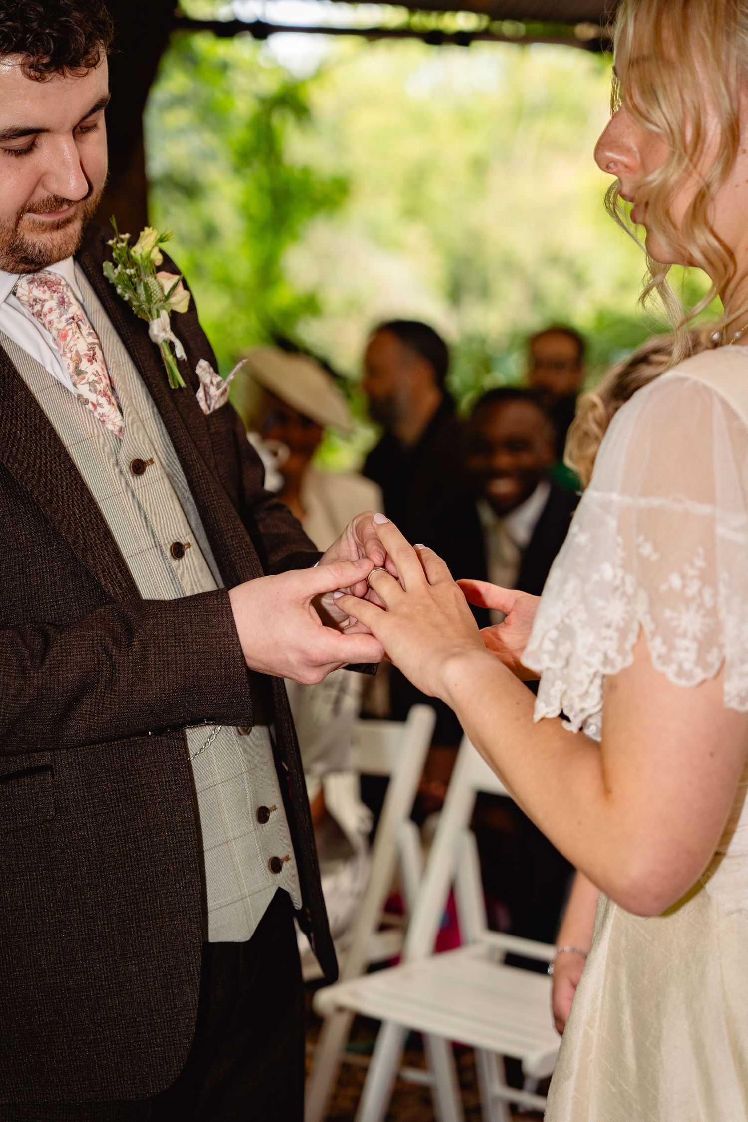 Groom places wedding ring on brides finger during outdoor wedding ceremony with guests seated behind.