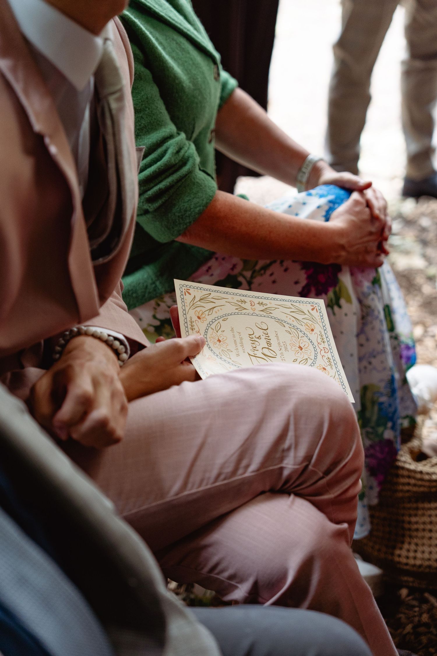Wedding guest in pink outfit holds decorative ceremony program booklet while seated next to guest in green cardigan.