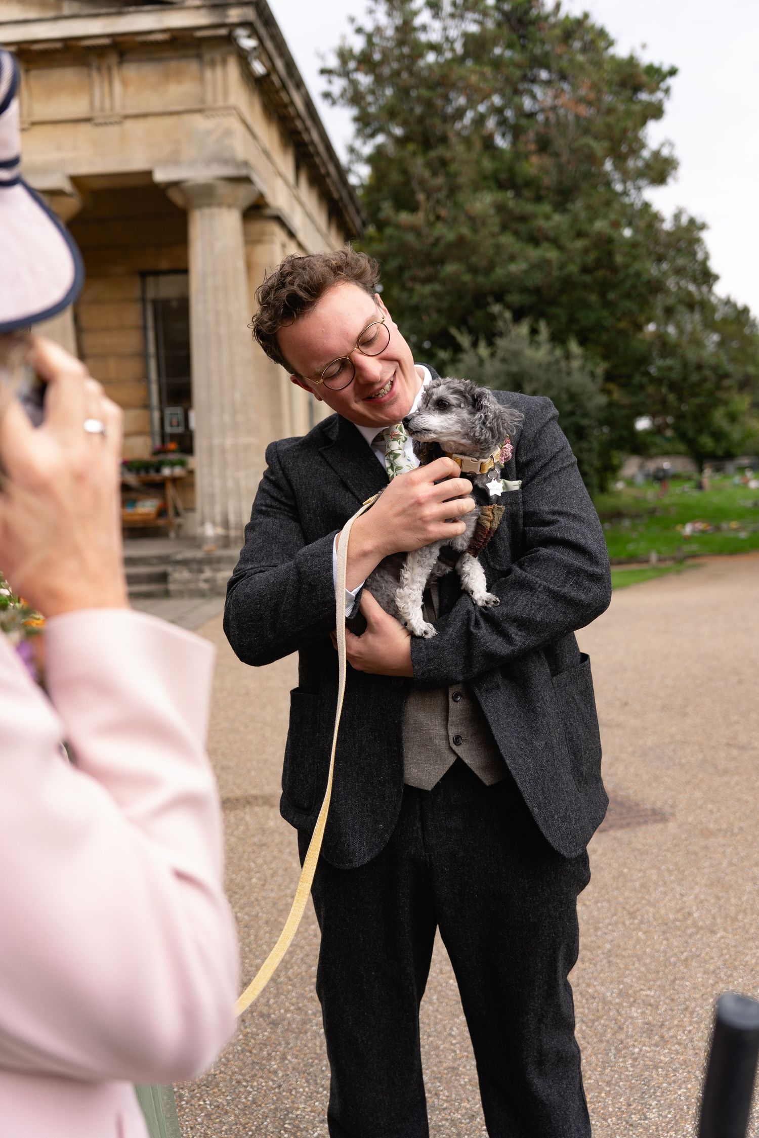Smiling groom in gray suit holds small dog with yellow leash at outdoor wedding venue.