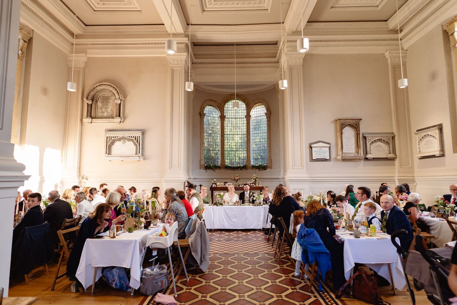 Elegant wedding reception in a grand historic hall with high ceilings, arched windows, and guests seated at white-clothed tables.