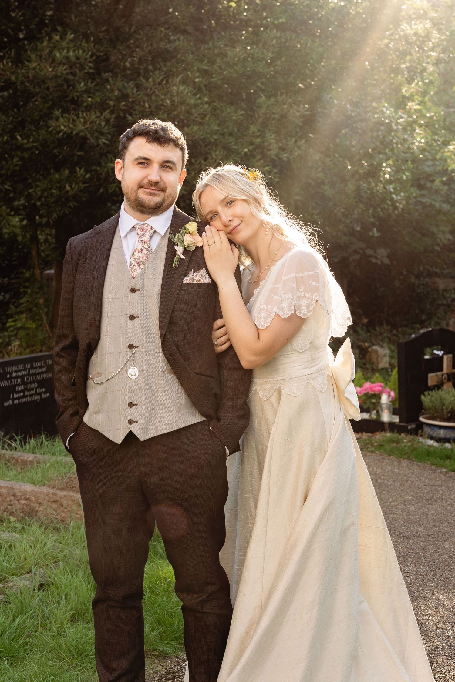 A bride and groom pose together outdoors in golden sunlight, she in a vintage cream gown, he in a brown suit with floral tie.