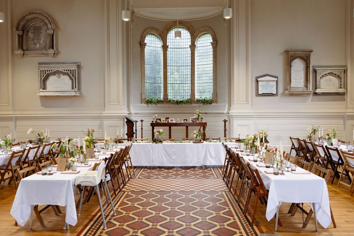 Elegant wedding reception hall with white-draped tables, wooden chairs, ornate arched window, and floral centerpieces on patterned rug.