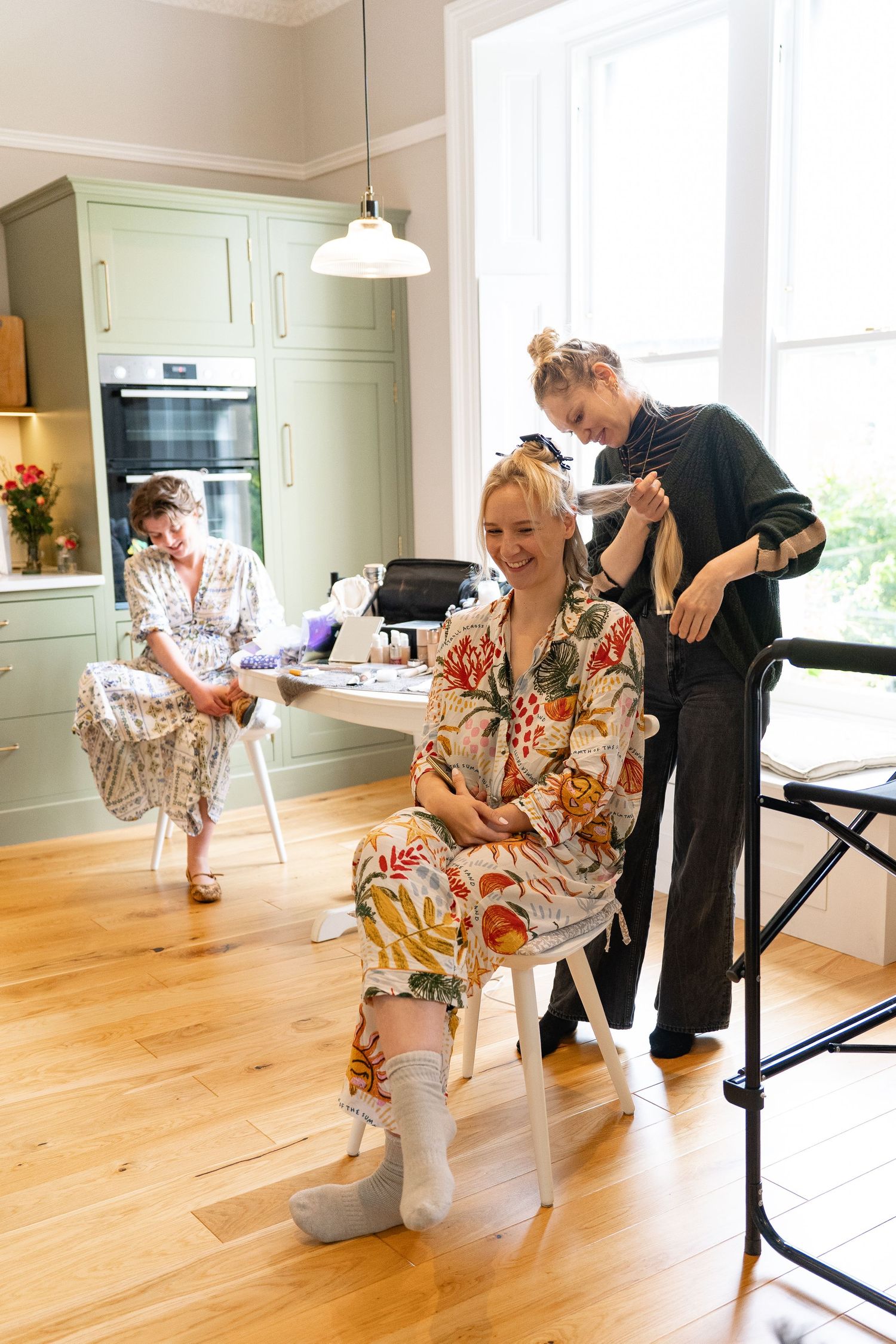 Woman in floral robe gets hair styled in modern kitchen while another woman sits in background.