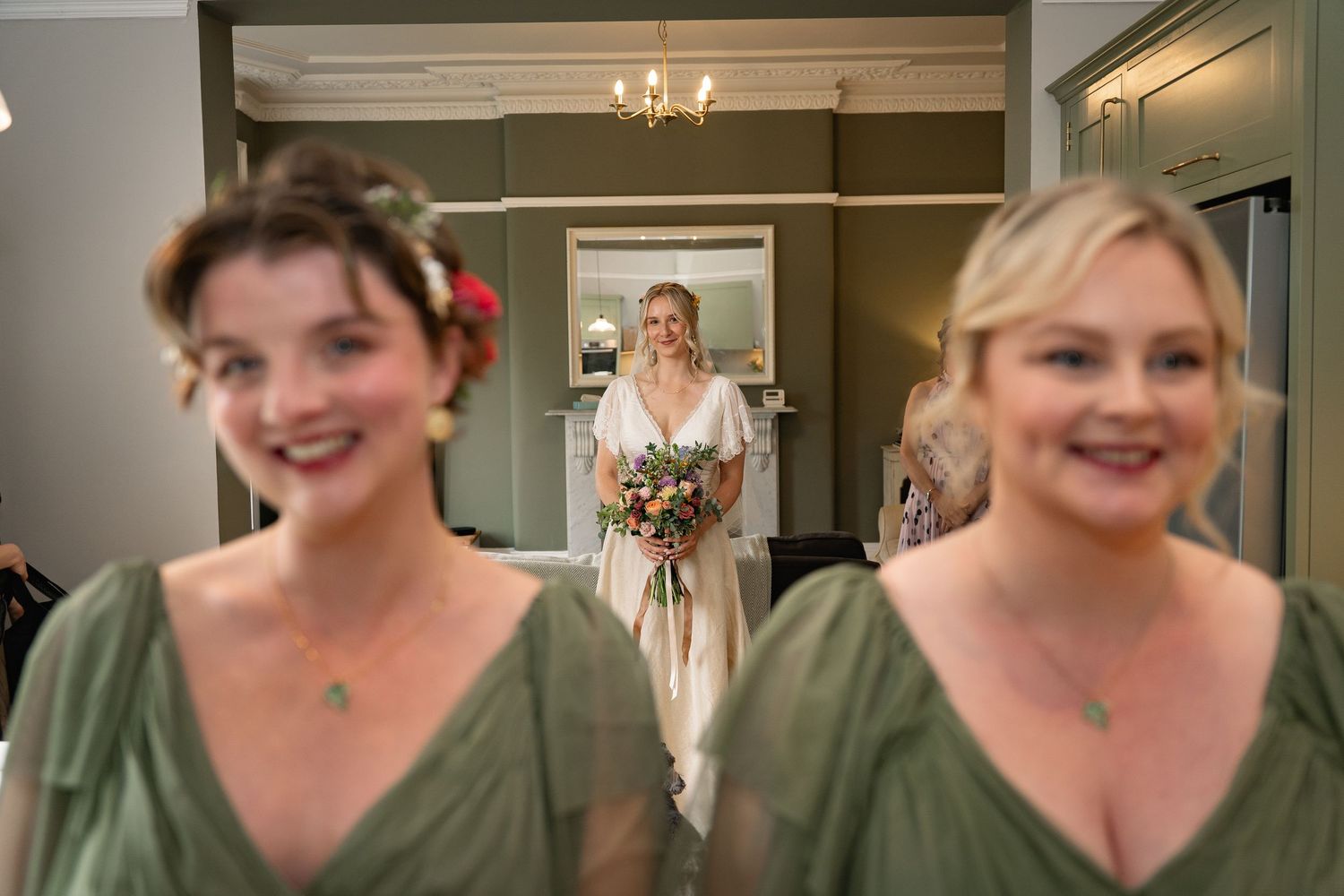 Bride in white gown holding bouquet stands between two smiling bridesmaids in sage green dresses inside elegant venue.