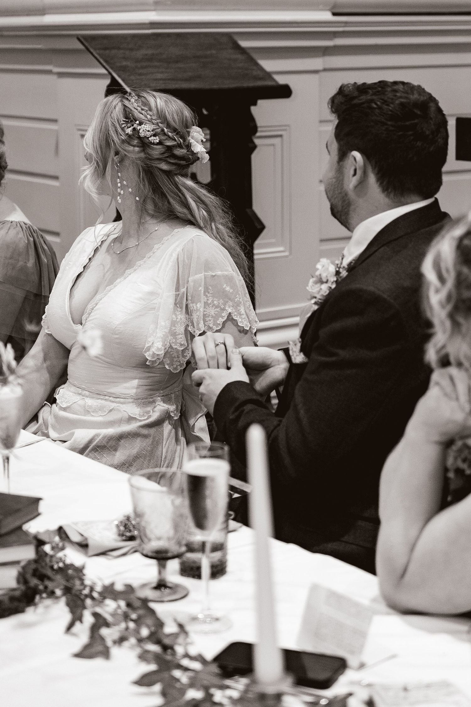 Black and white photo of a bride and groom holding hands at their wedding reception table, surrounded by guests and floral decor.