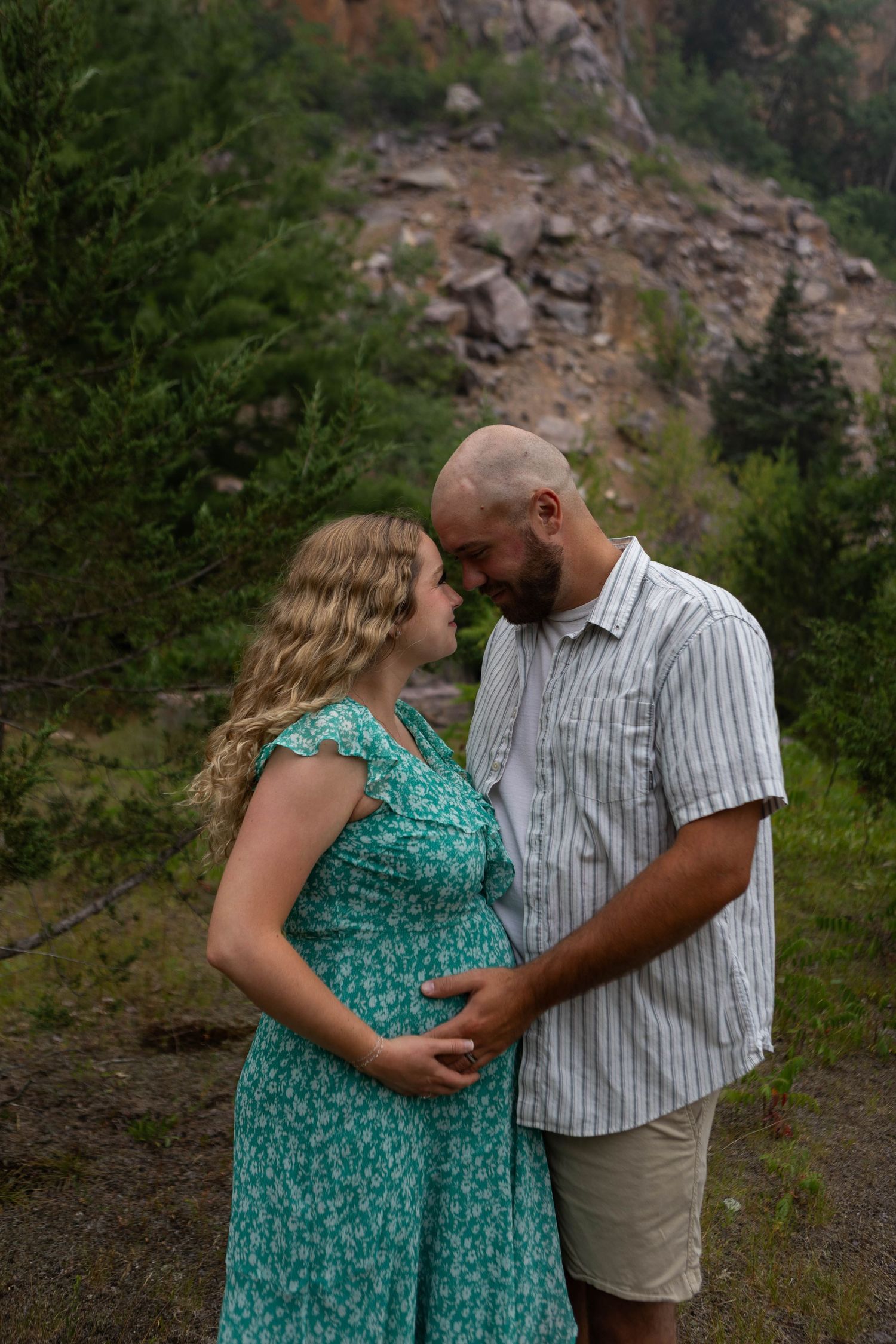 Pregnant couple tenderly touching foreheads outdoors, woman in teal floral dress, man in striped shirt, rocky mountain backdrop.