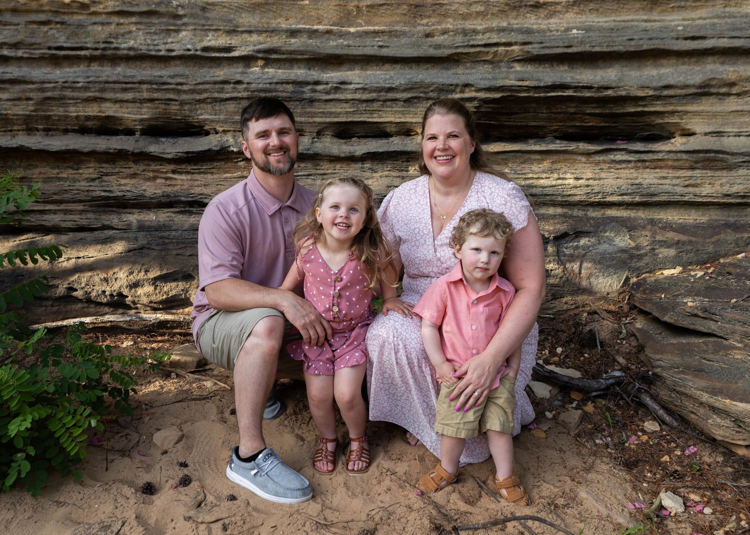 Family of four smiling in front of a stone wall outdoors, parents with two young children in pink and coral outfits.