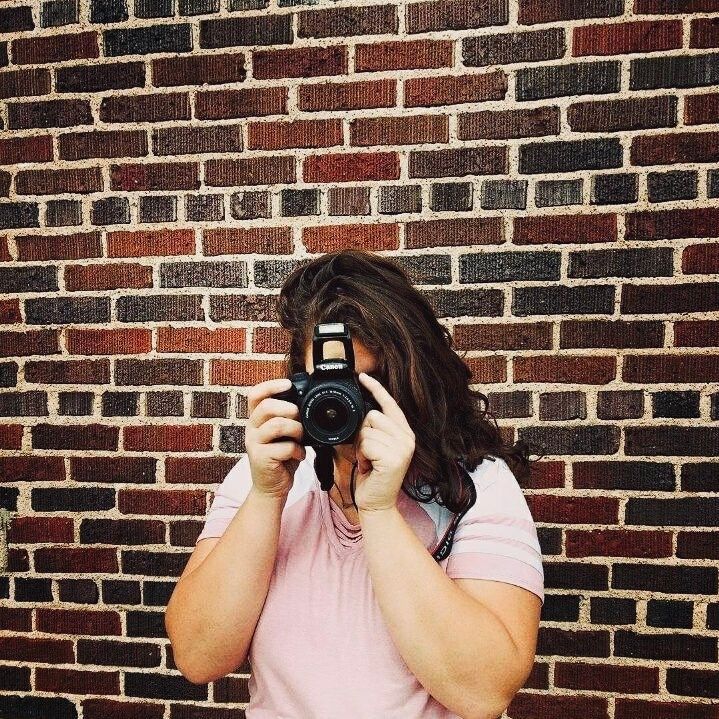 Person wearing pink takes photos in front of textured brick wall. Mauston photographer.