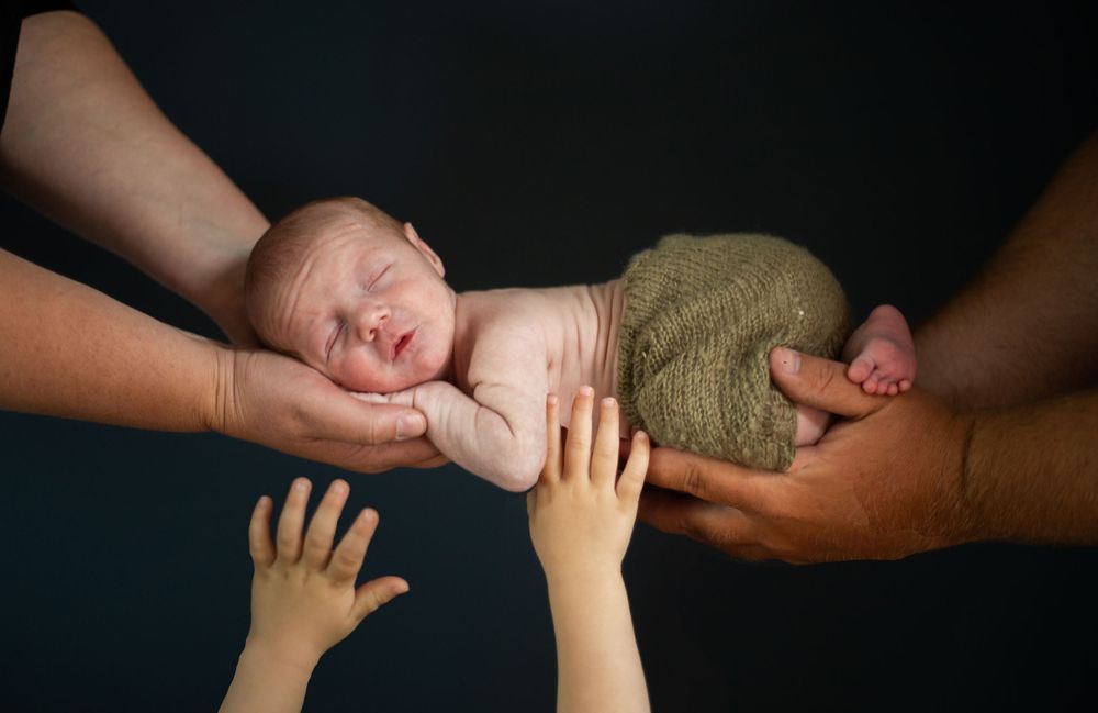 Multiple hands gently supporting sleeping newborn wrapped in olive fabric. Mauston photographer.