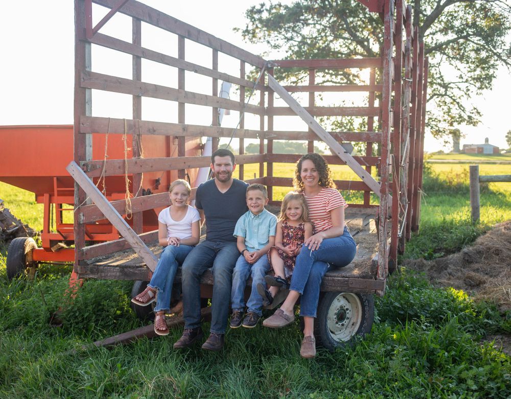 Group sitting on farm equipment near a red barn structure during sunset. Mauston photographer.
