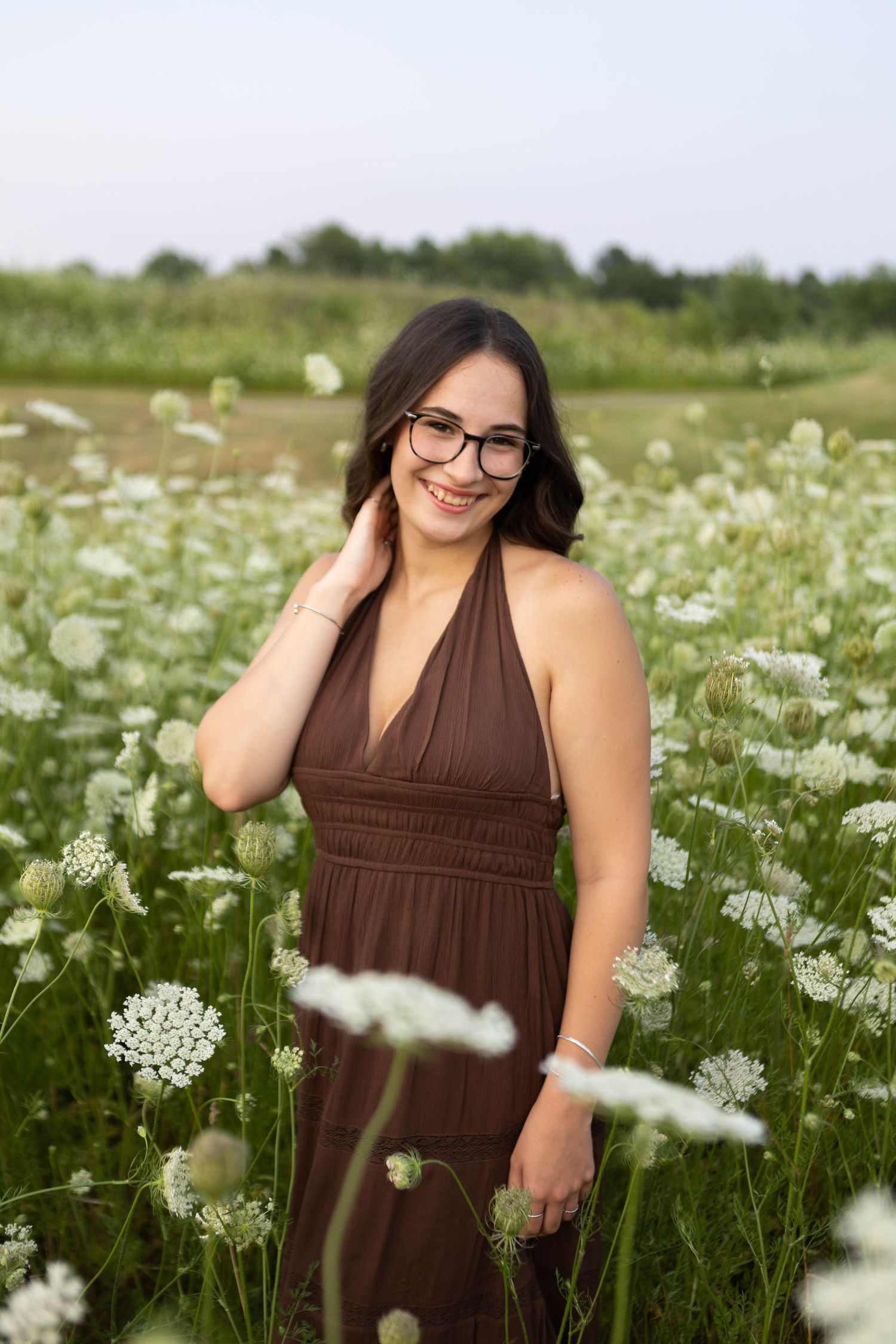 Young woman in brown halter dress and glasses smiling in a field of white wildflowers on a cloudy day.