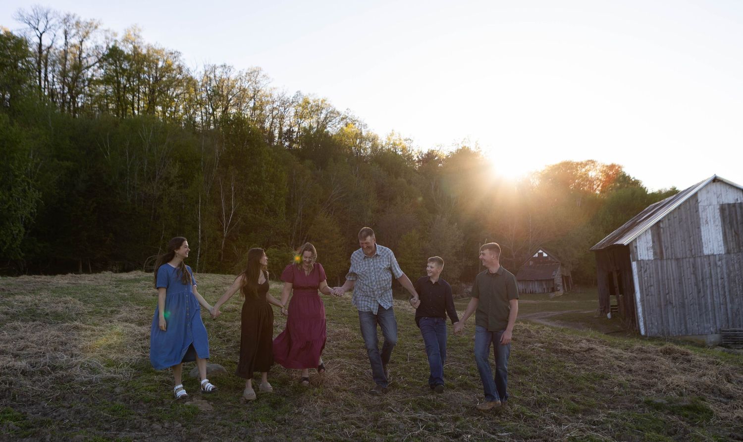 Family of six walks hand-in-hand across a sunlit farm field at golden hour with trees and barn in background.