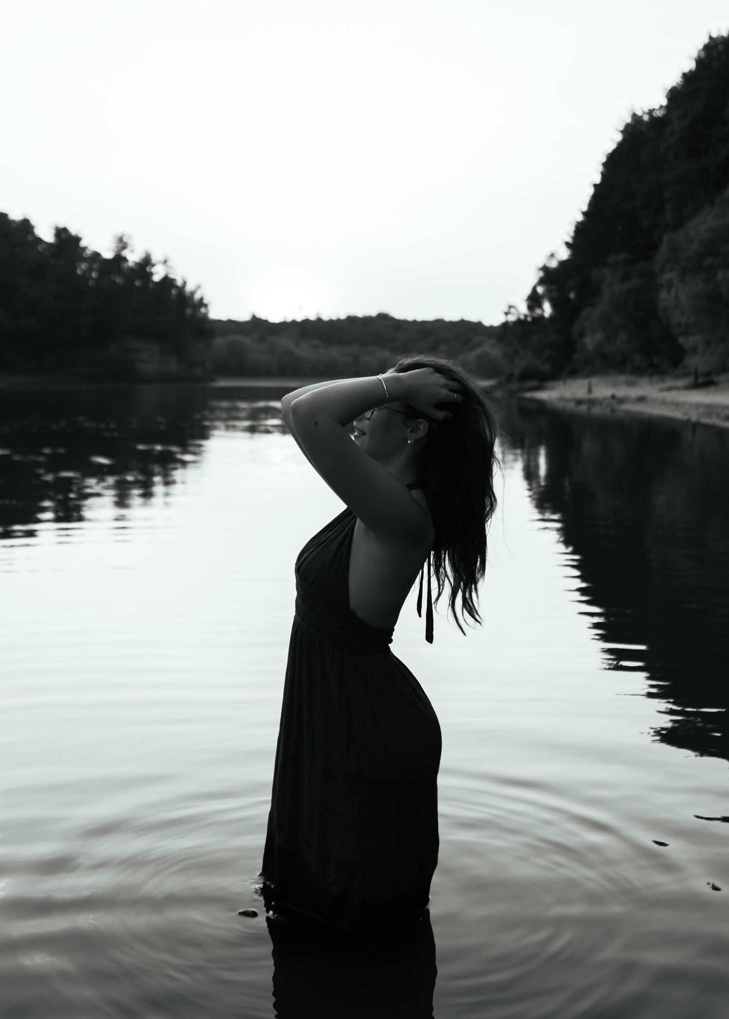 Black and white photo of a woman standing in a river, hand in hair, surrounded by trees and cliffs.