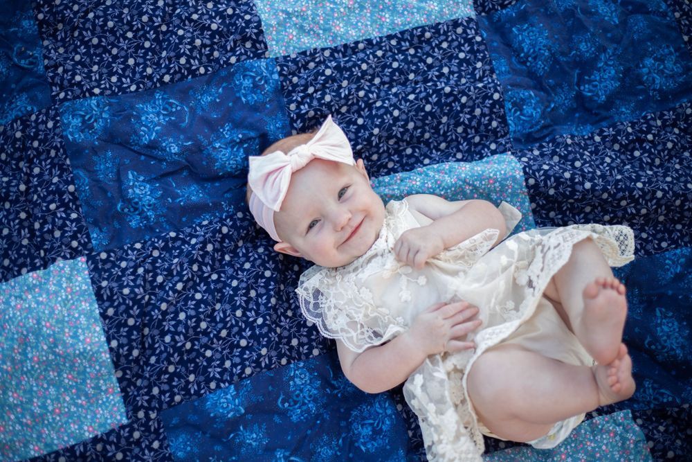 Baby in white lace dress lying on blue patchwork quilt. Mauston photographer.