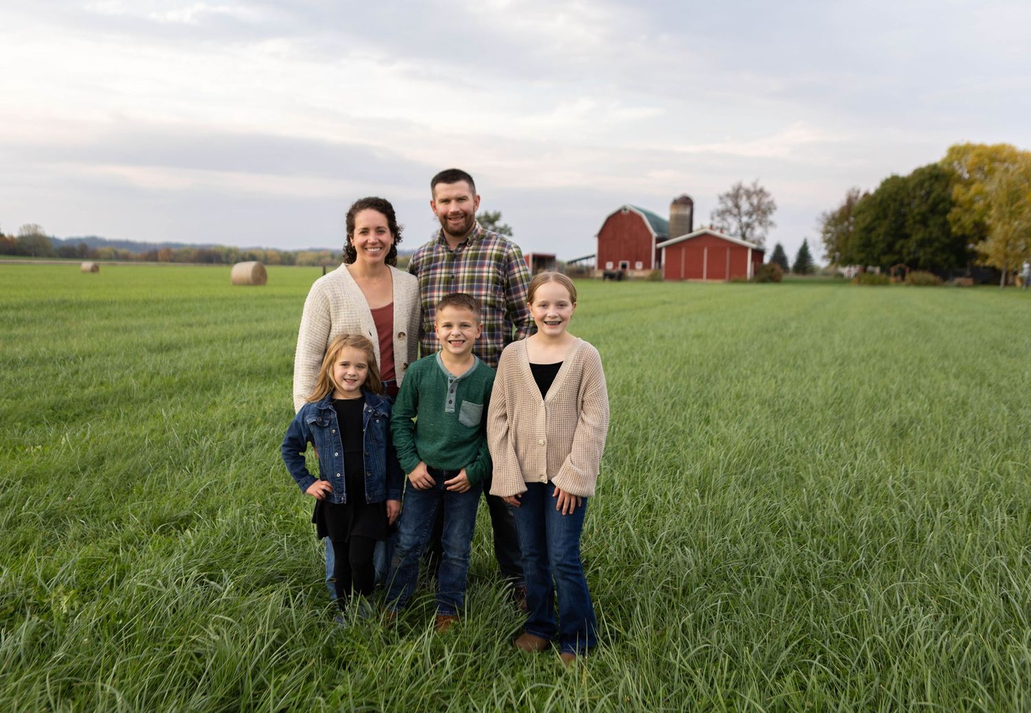 Family portrait in a vast green field with red barn and rural landscape in background. Mauston photographer.