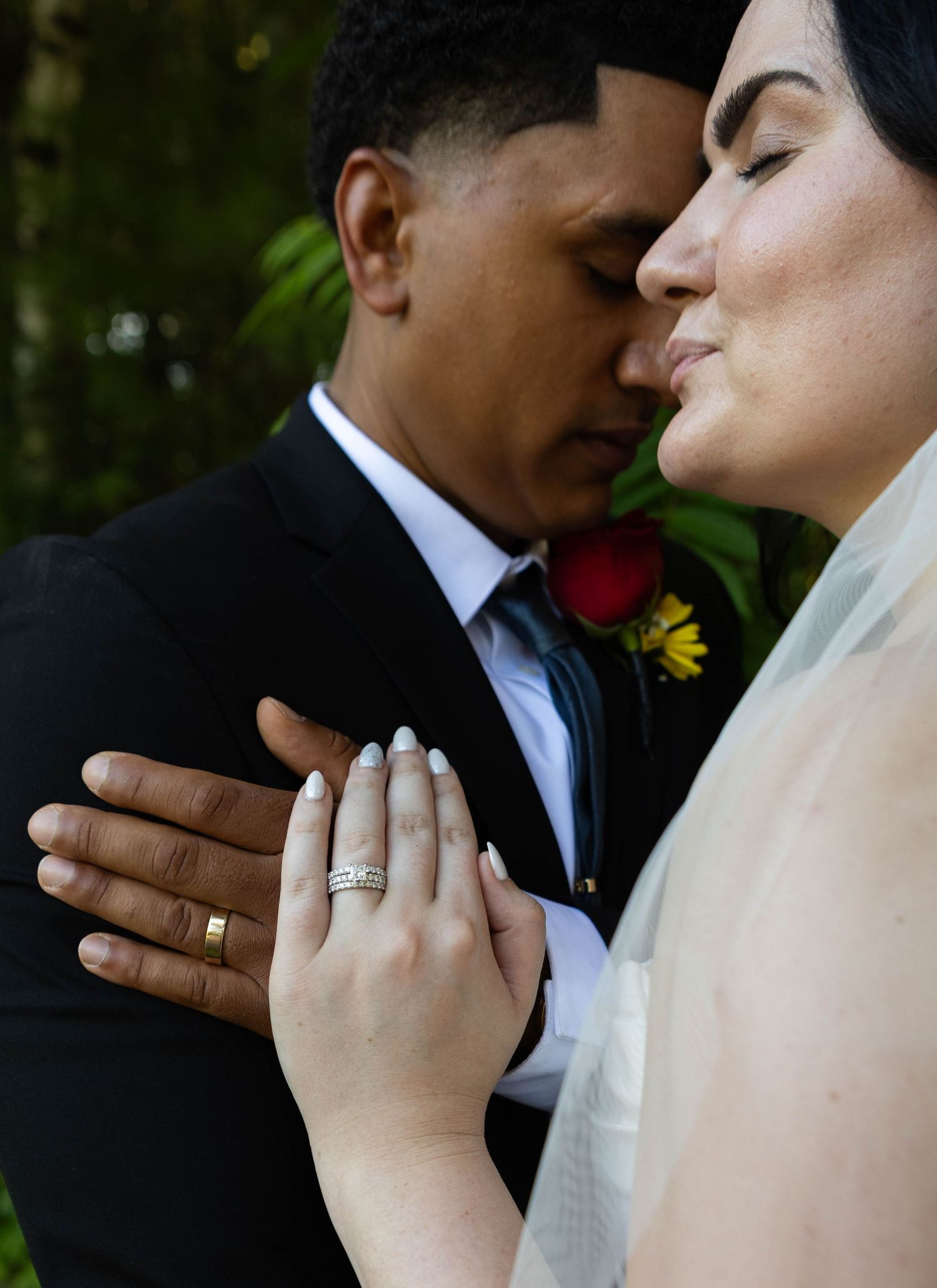 Newlywed couple in wedding attire displaying their gold and diamond rings, sharing an intimate moment outdoors.