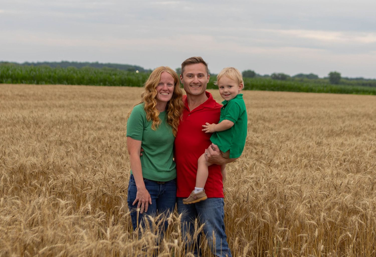 A smiling family of three poses in a golden wheat field, with the man holding a young child, all wearing green and red shirts.