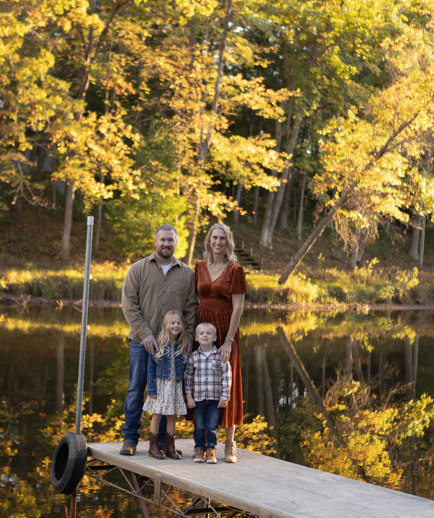 Family portrait on wooden dock during autumn with golden foliage reflecting in lake. Mauston photographer.