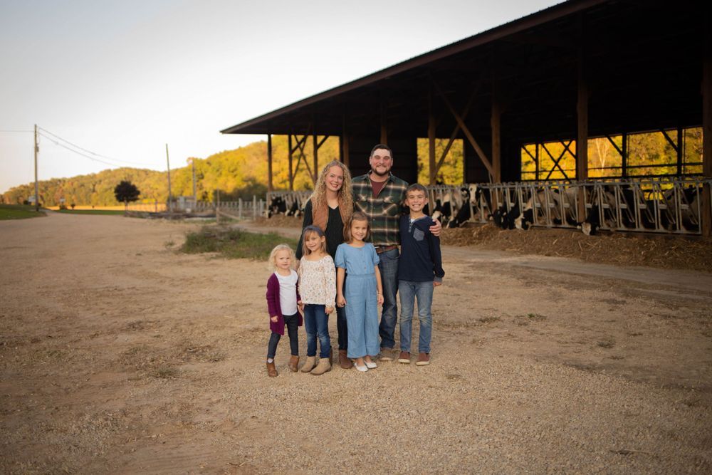 People gathered outside a barn at sunset with farm equipment visible in the background. Mauston photographer.