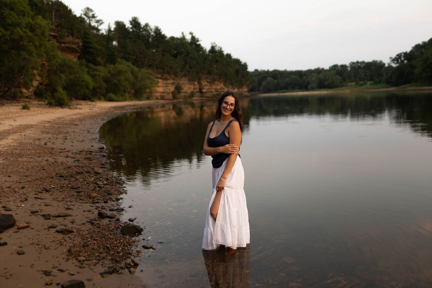 Woman in white skirt standing in shallow river water near sandy shore with red rock cliffs and trees.