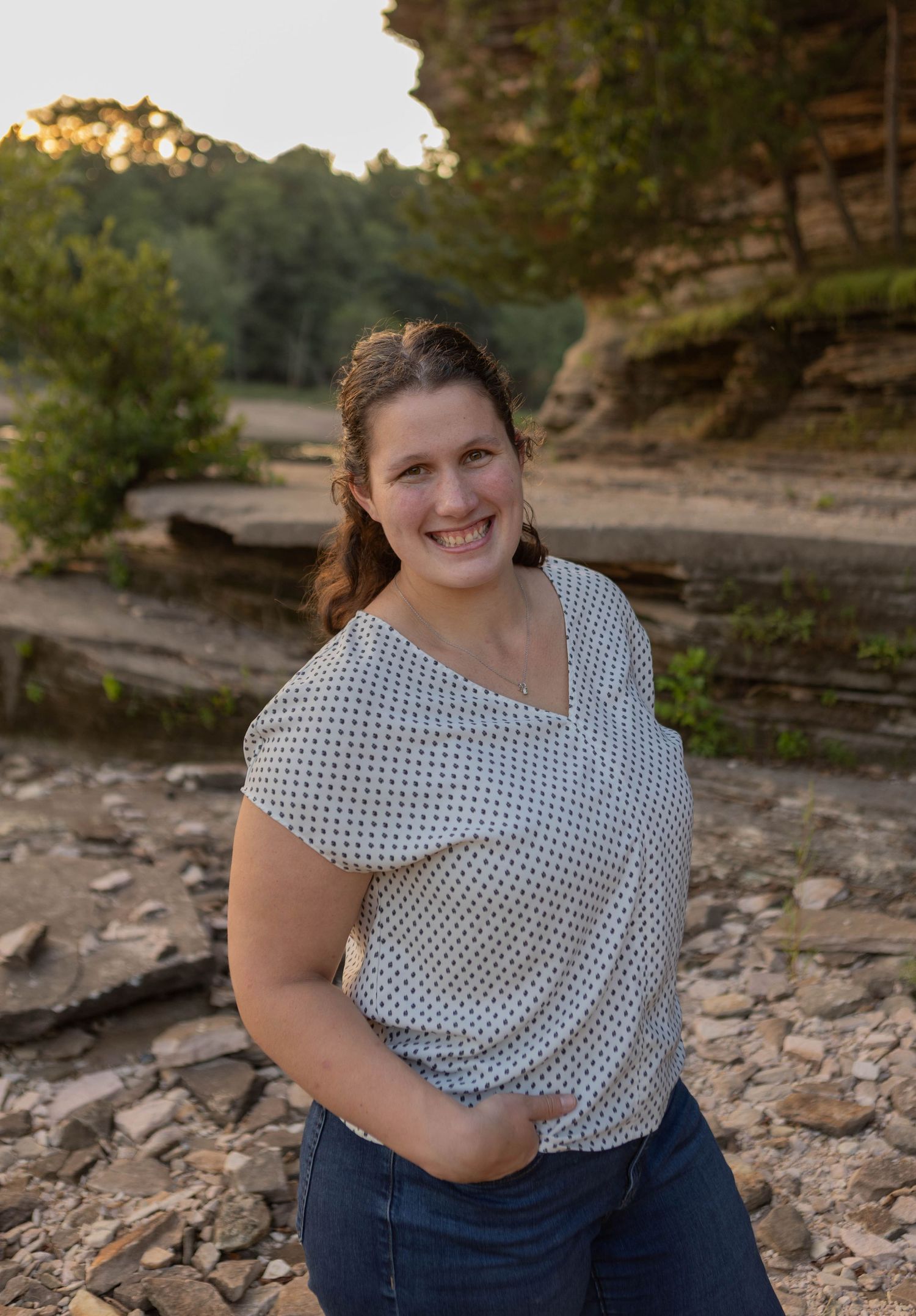 Smiling woman in polka dot top and jeans poses outdoors near rocky cliffs and trees at golden hour.