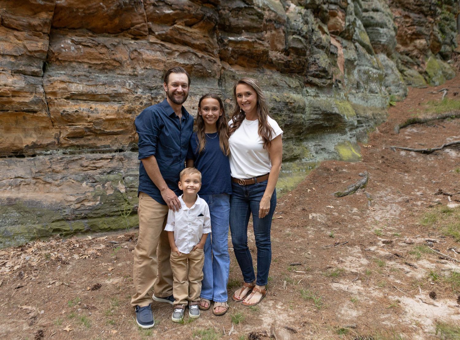 Family of four posing together in front of a red sandstone rock wall on a nature trail.