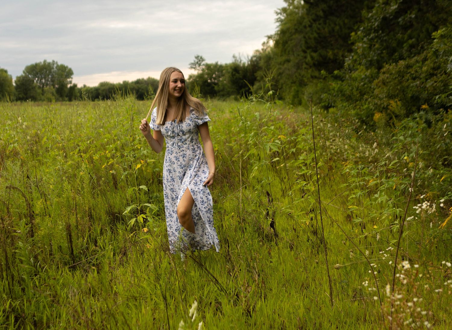Figure in floral dress walking through tall grass in summer meadow. Mauston photographer.