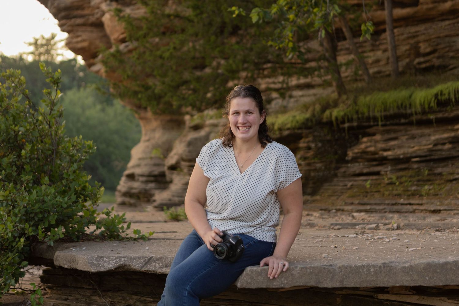 Woman smiling and sitting on a rock ledge beneath a sandstone cliff in a scenic outdoor nature setting.