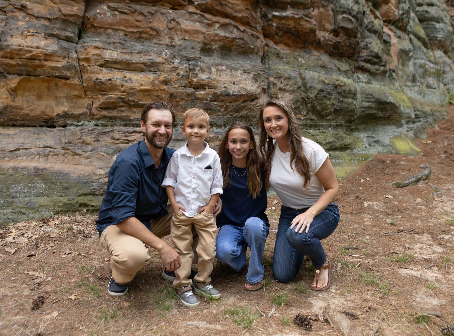 Family of four smiling together outdoors near a rocky sandstone cliff, posing for a portrait photo.