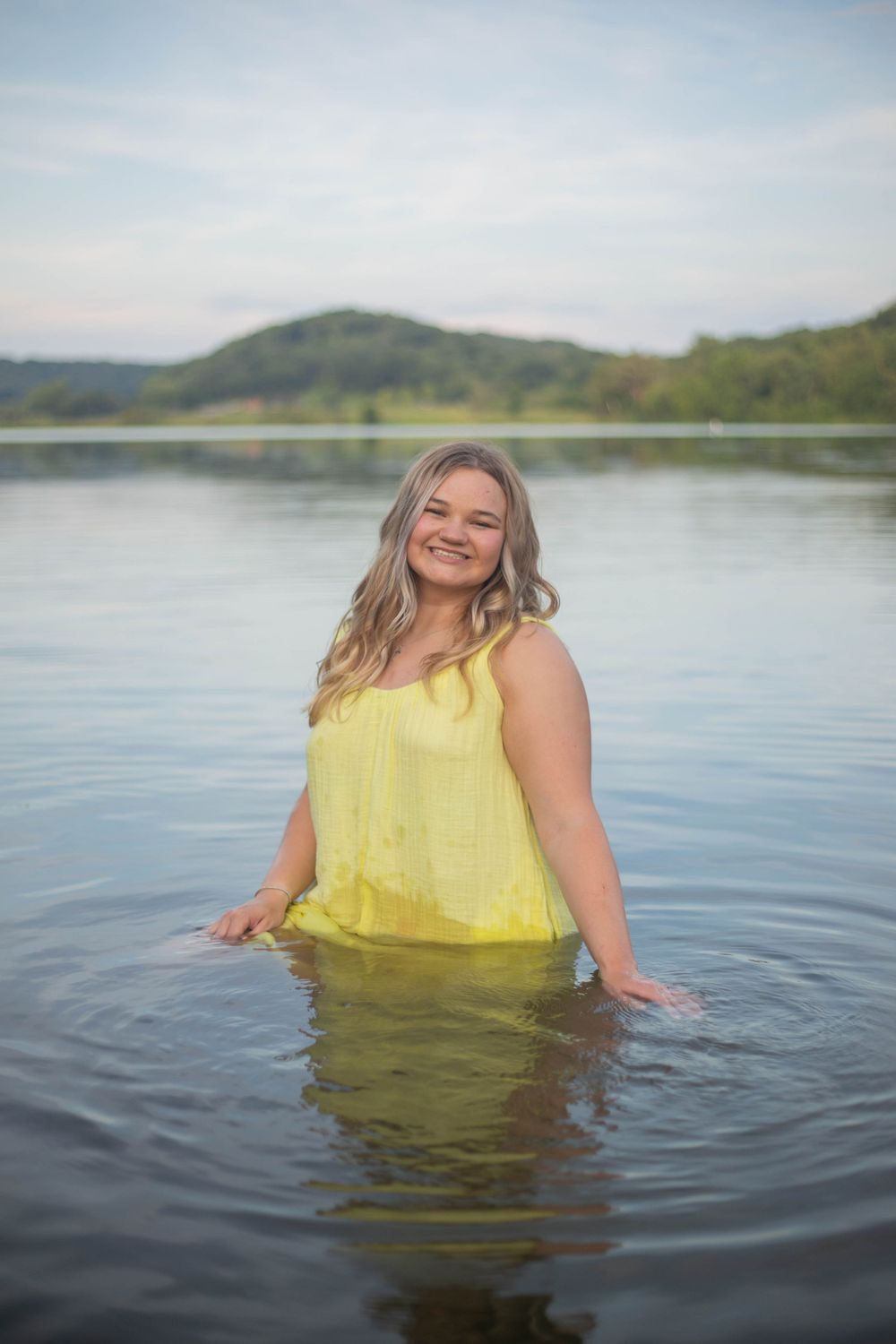 Person in yellow dress wading in calm lake water with mountains in background. Mauston photographer.