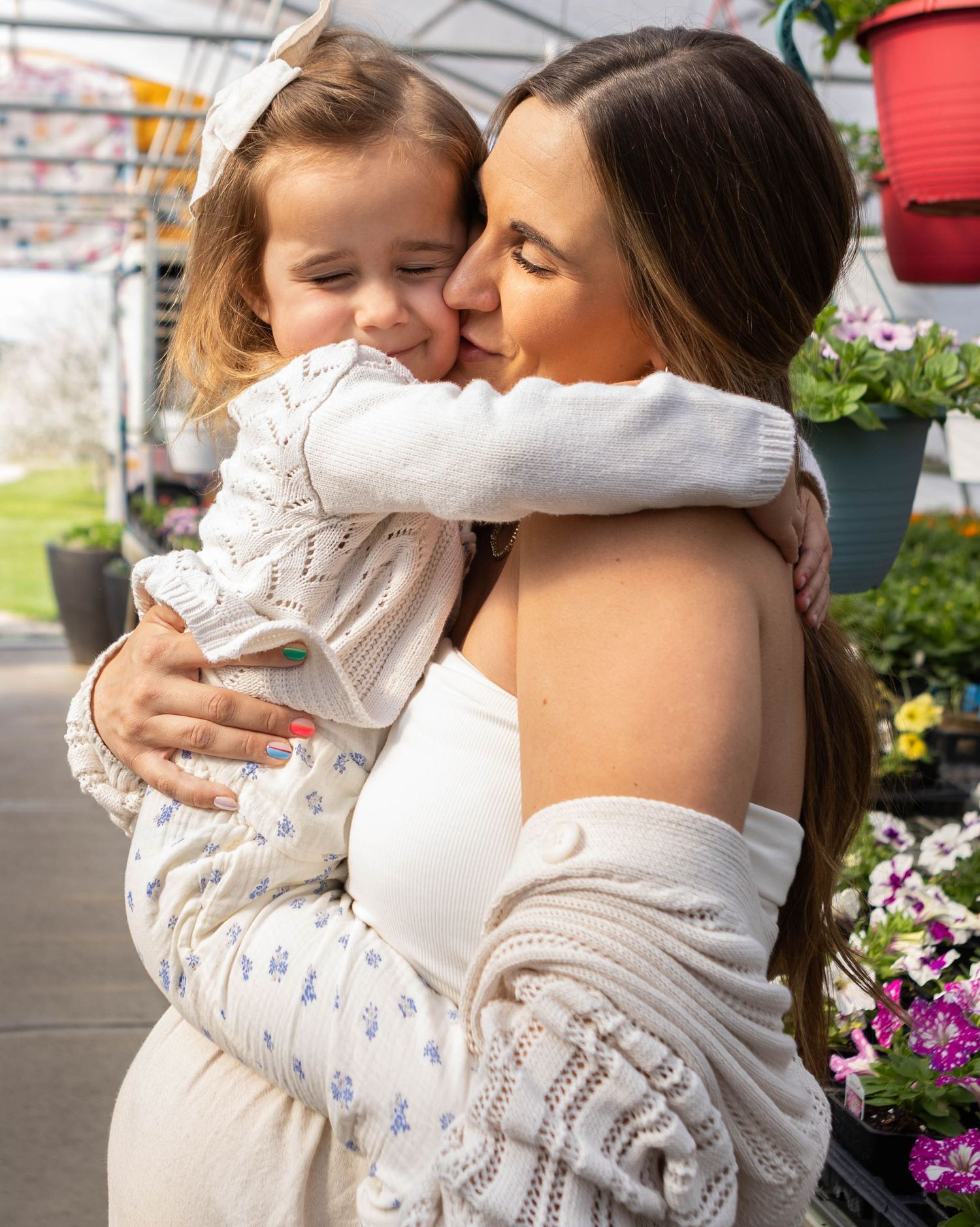 A mother kisses her young daughter while embracing her in a flower greenhouse, both wearing white outfits.