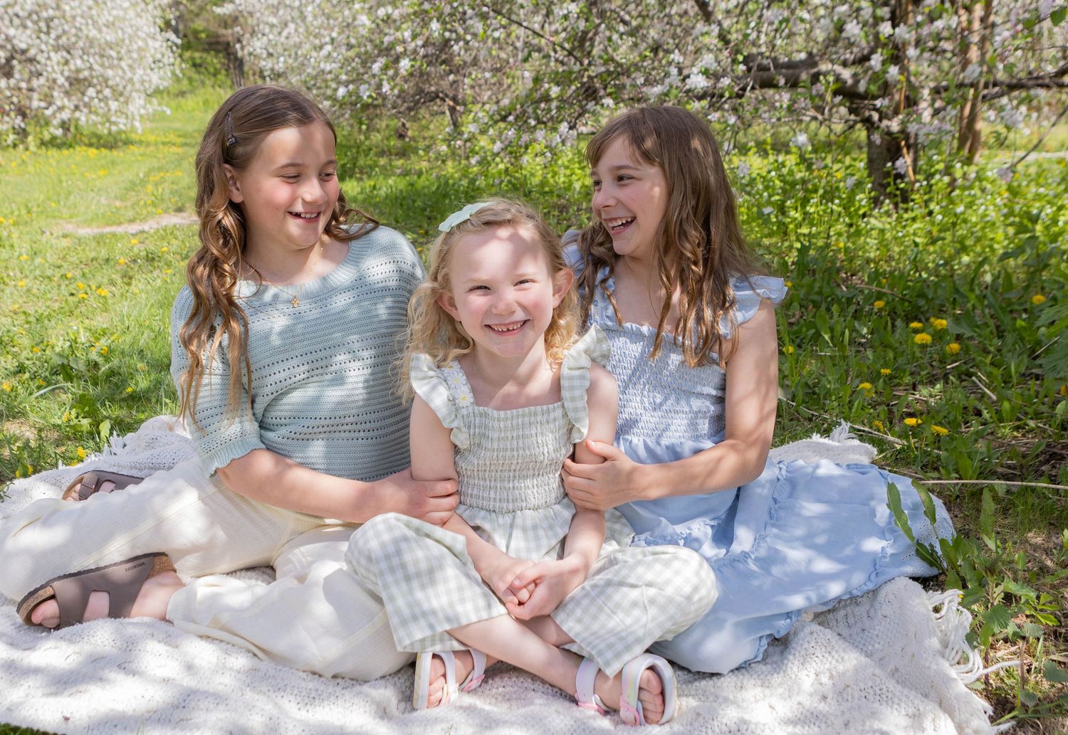 Three smiling girls in pastel dresses sitting on a blanket outdoors among blooming spring trees.