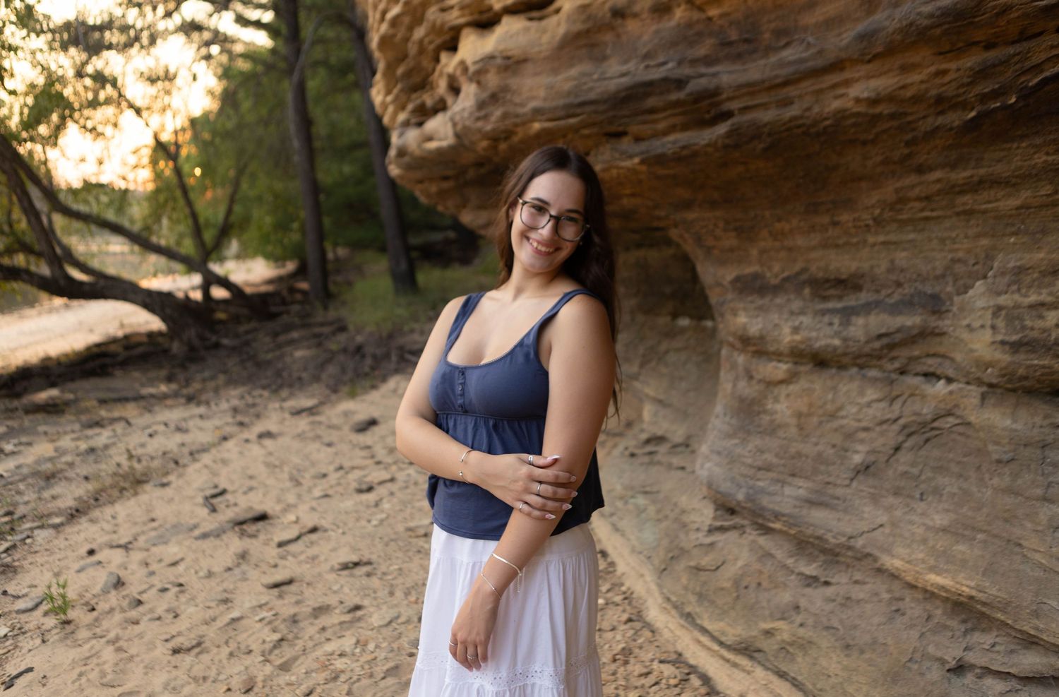 Young woman in glasses and white skirt smiles beside a sandstone rock formation in a wooded outdoor setting.