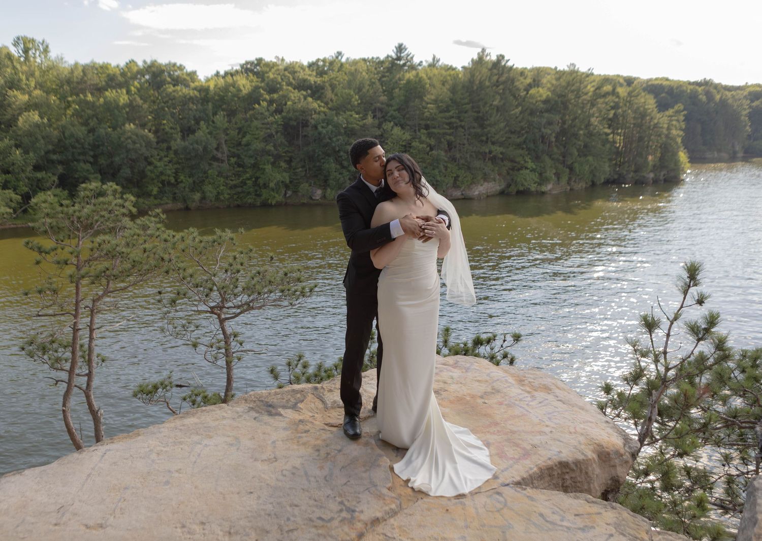 Bride and groom embrace on rocky cliff overlooking scenic river and lush forest during outdoor wedding photo session.
