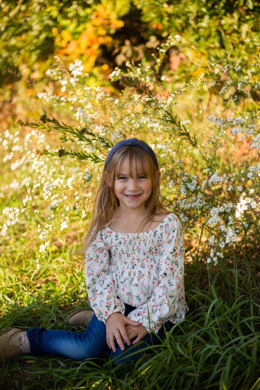 Young child sitting in tall grass with flowering trees and golden sunlight in background. Mauston photographer.
