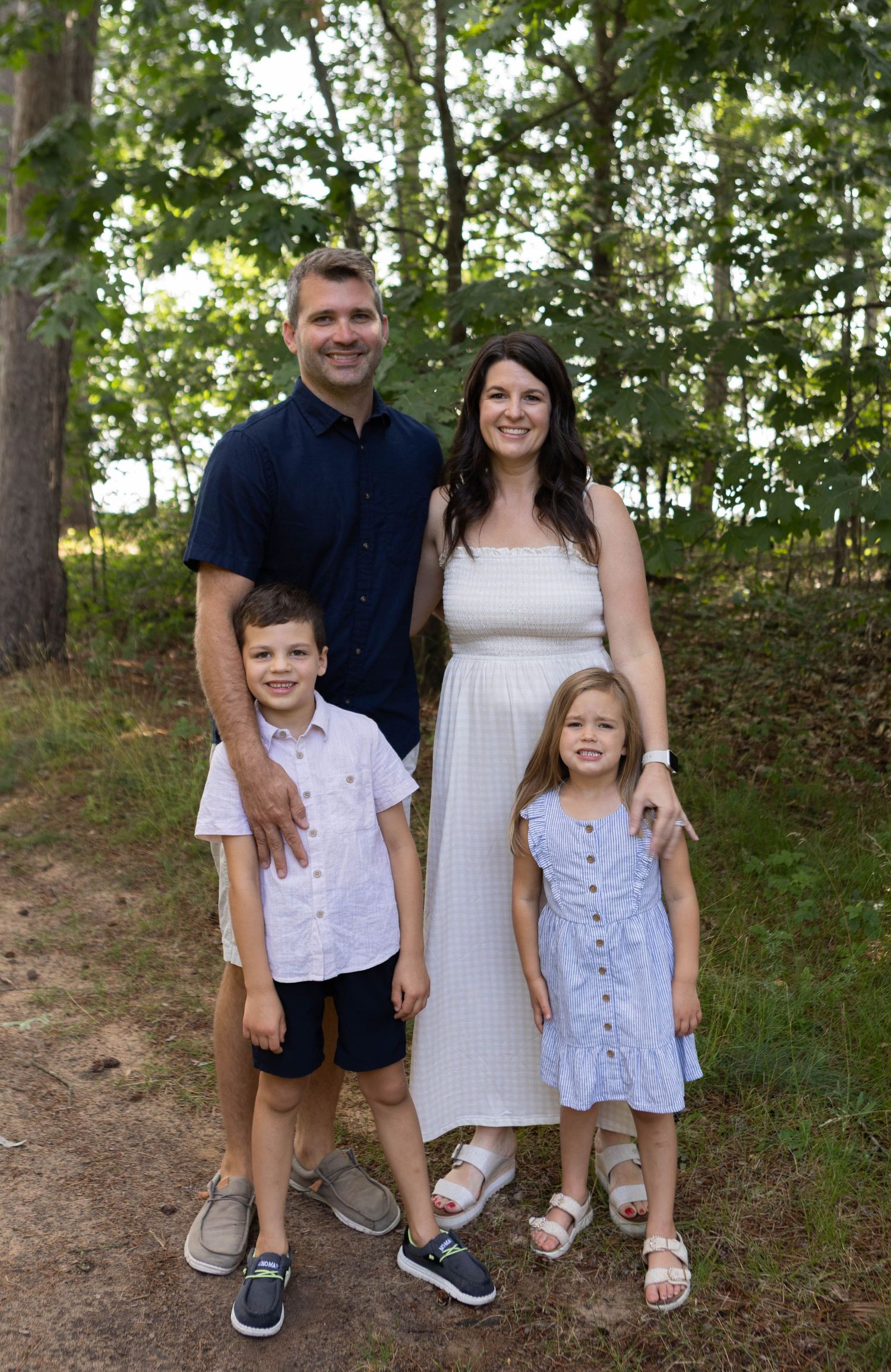 A happy family of four poses on a wooded trail, parents with two young children dressed in coordinated outfits.