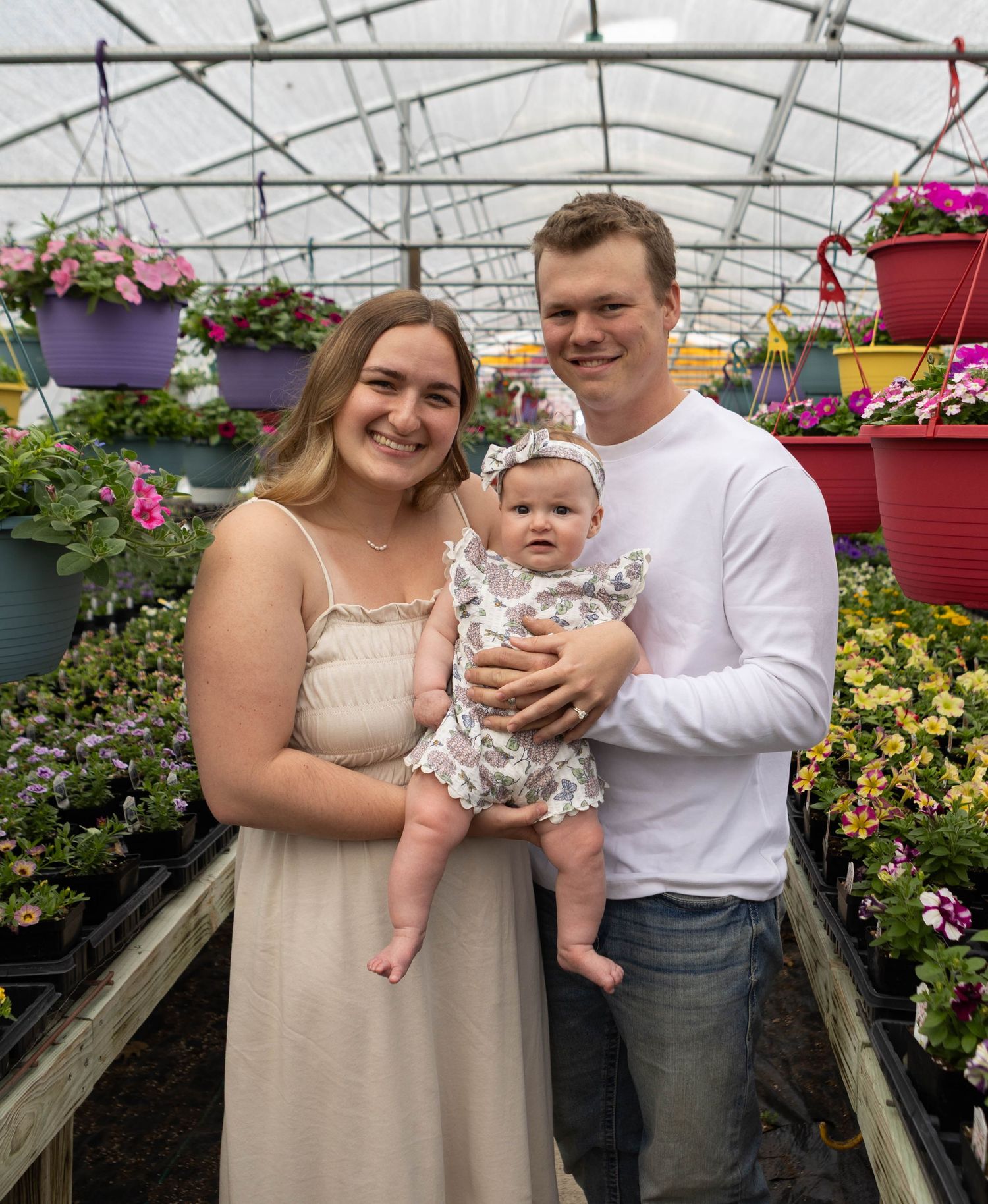 A young family smiles together in a colorful greenhouse, with parents holding their baby girl among vibrant hanging flower baskets.