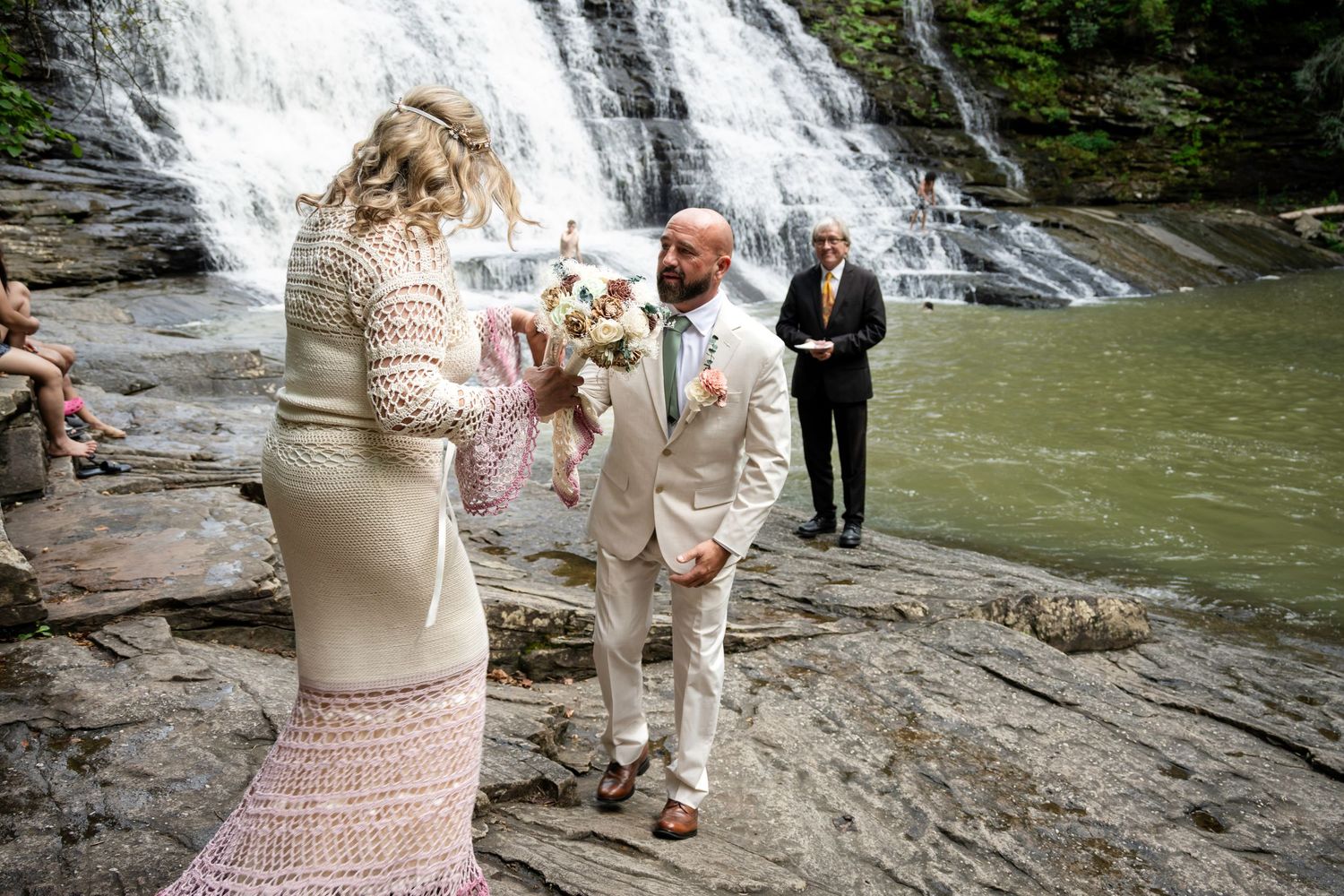 Couple exchanging vows at waterfall wedding ceremony on rocky terrain with officiant and guests in background.