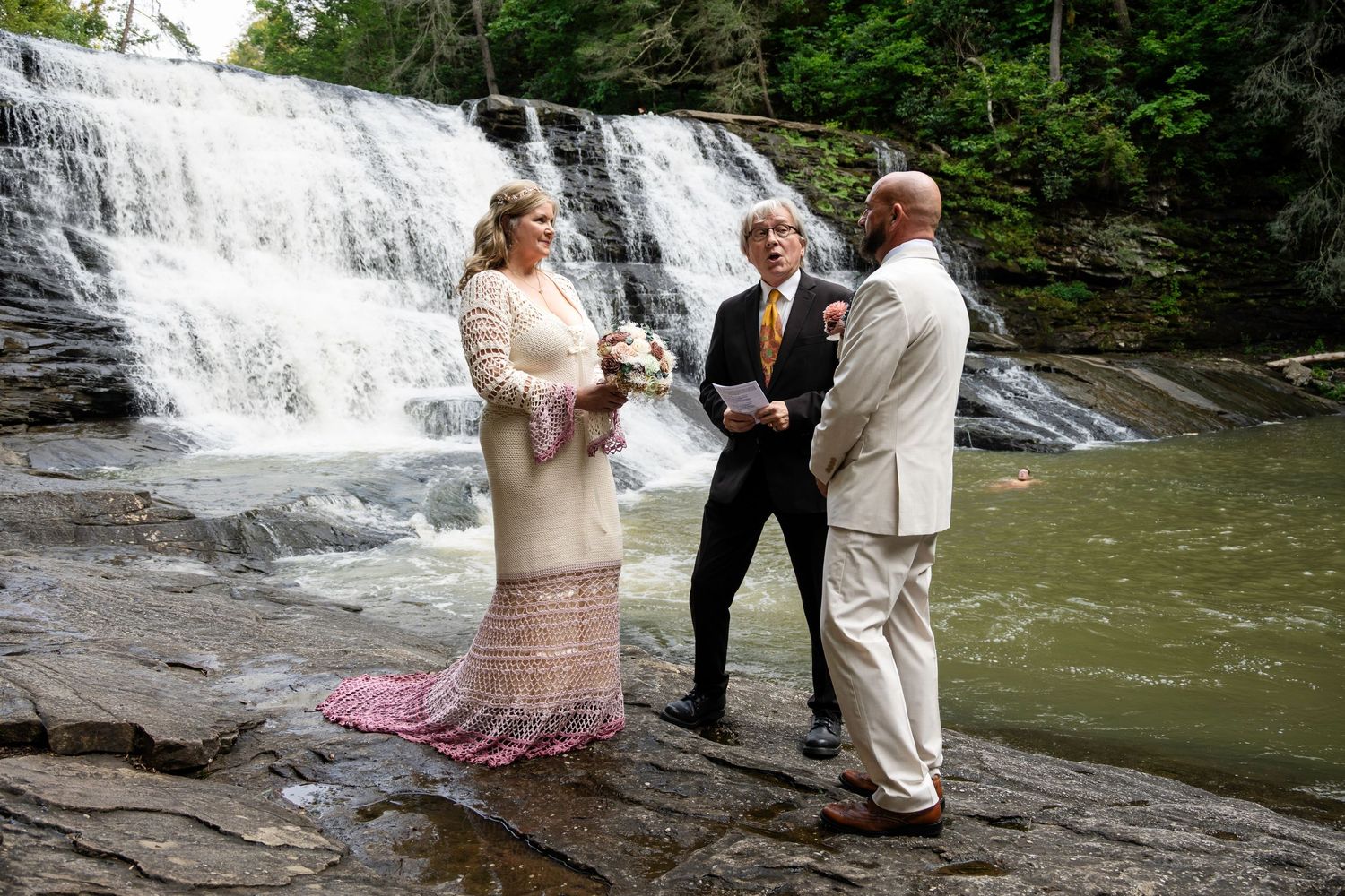 Couple exchanging vows at a waterfall wedding ceremony outdoors, officiant holding papers, bride in lace dress.