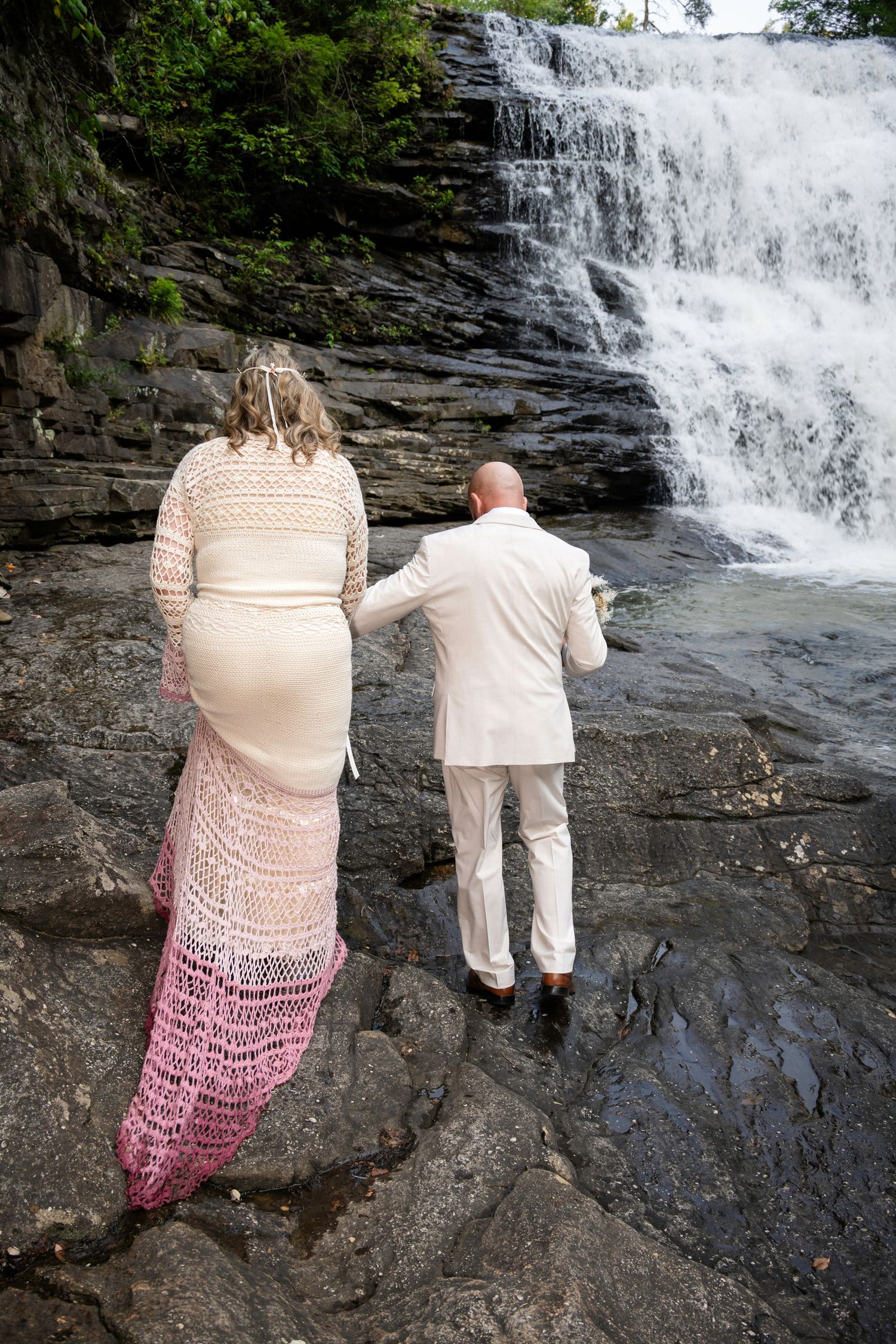 Bride and groom stand on rocks before a waterfall, she in pink crochet dress, he in white suit.