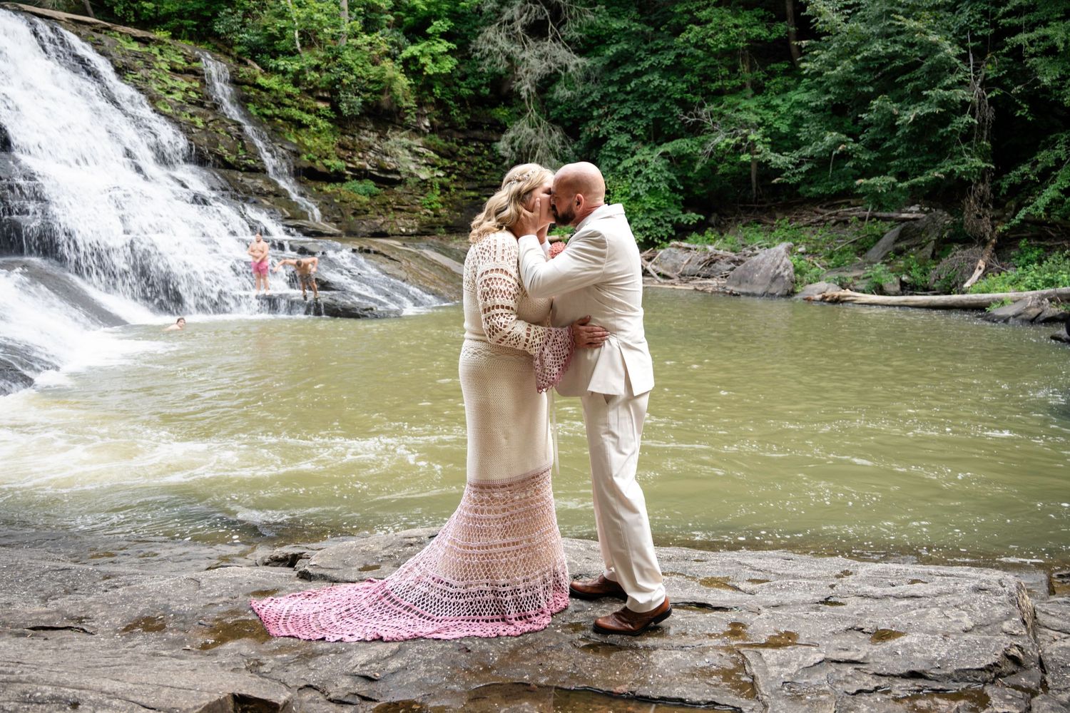 Couple sharing a romantic kiss in wedding attire beside a scenic waterfall on rocky terrain surrounded by lush greenery.