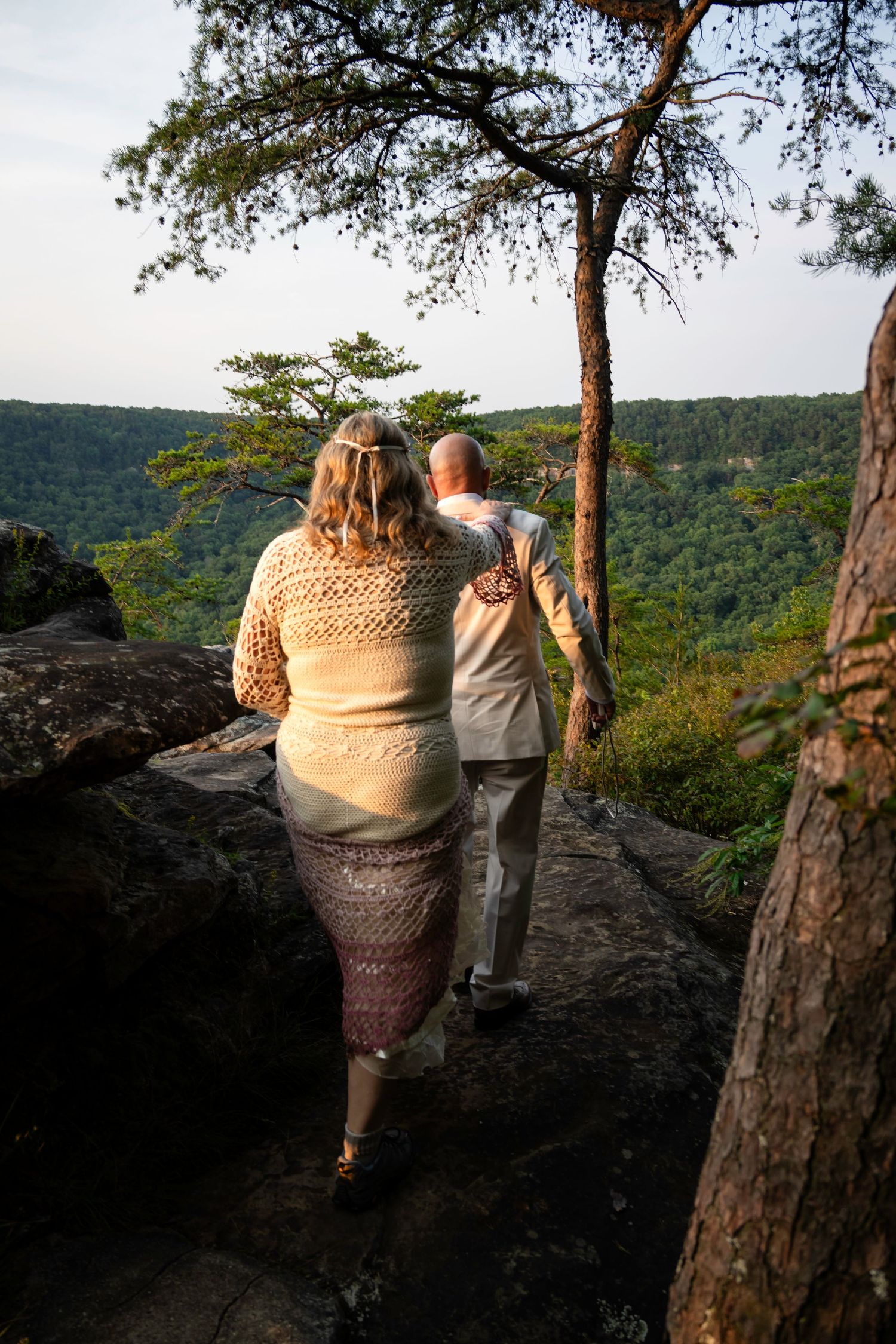 Couple standing on rocky overlook admiring lush green valley views, woman in knit sweater and skirt, man in light outfit.