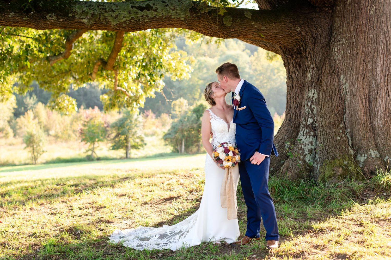Bride and groom kissing under a large oak tree in a sunlit outdoor setting, bride holding a colorful bouquet.