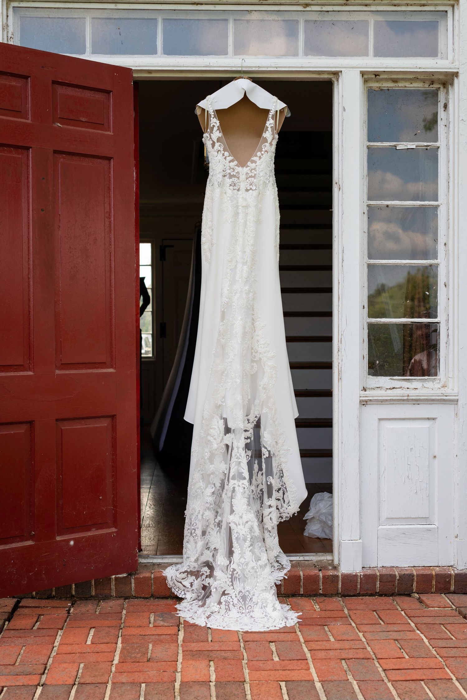 White lace wedding dress with train hanging on a doorframe between red and white doors on a brick porch.