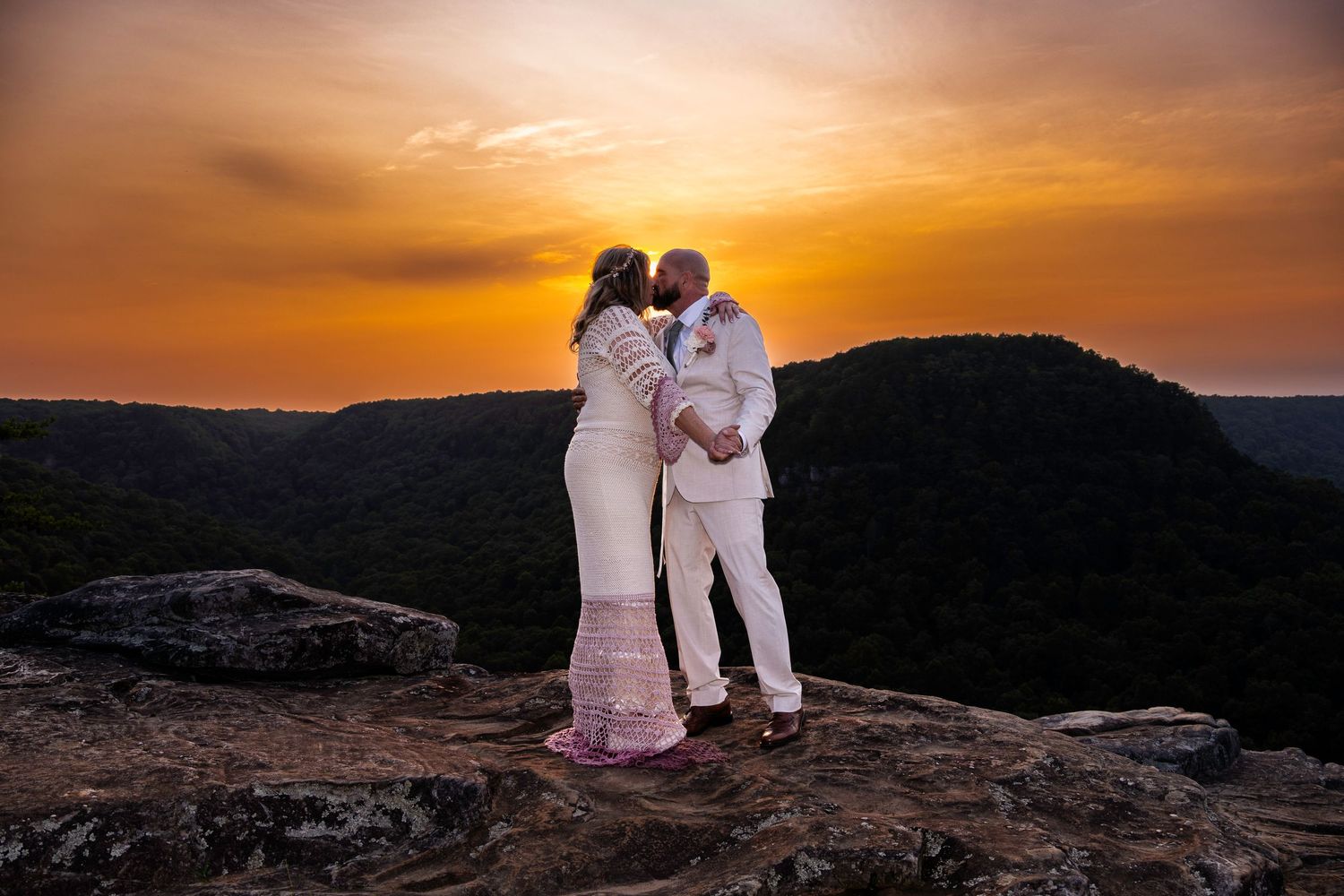 Couple sharing a romantic kiss at sunset on a rocky cliff overlooking lush green mountains in a stunning outdoor wedding.