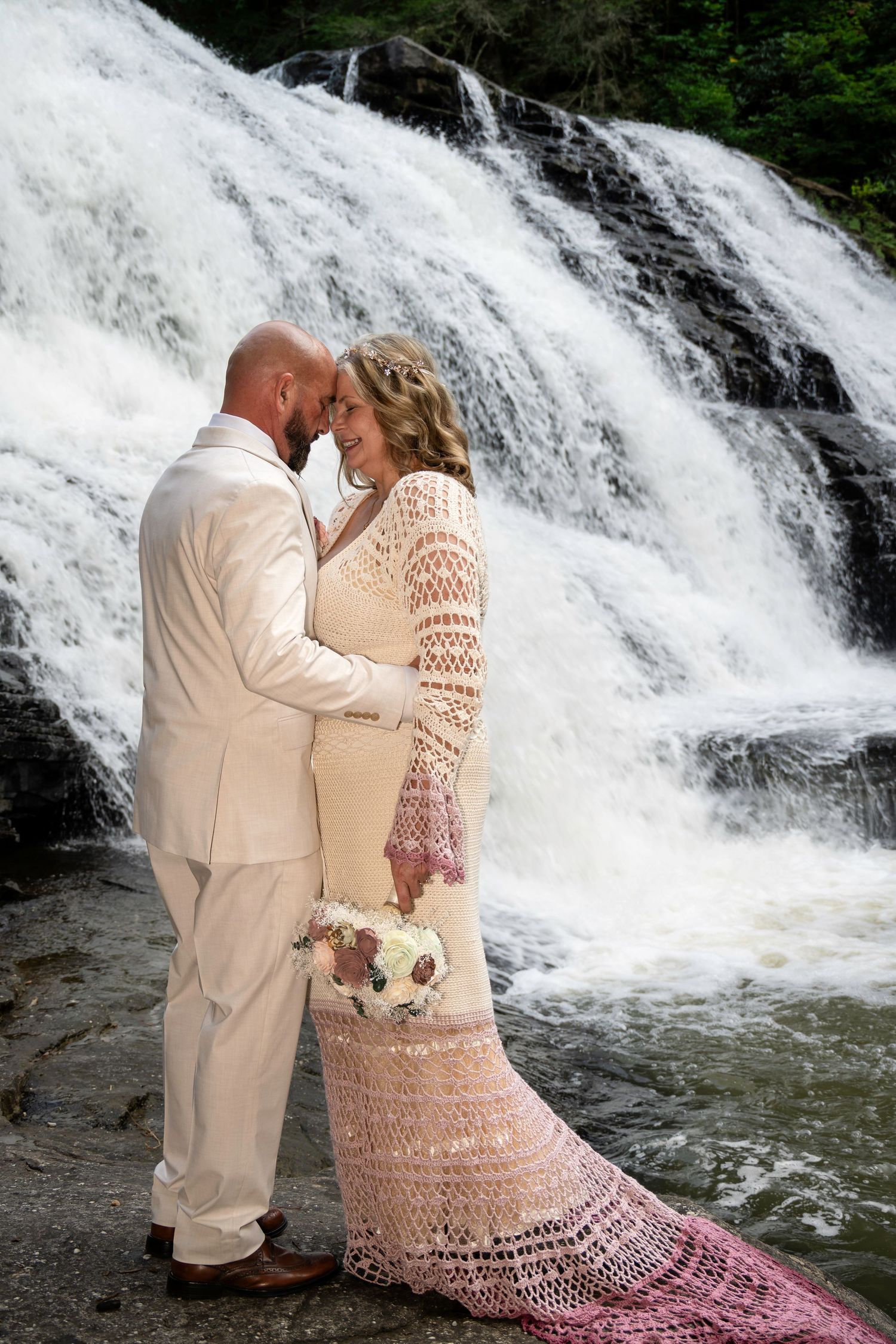 Bride and groom embrace before a stunning waterfall, she wearing a pink crochet dress holding a bouquet, he in a white suit.