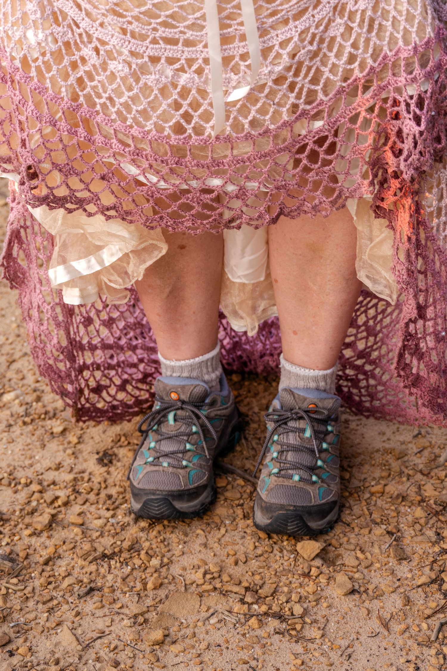 Person wearing hiking shoes and socks under a pink crochet lace skirt standing on dirt ground.