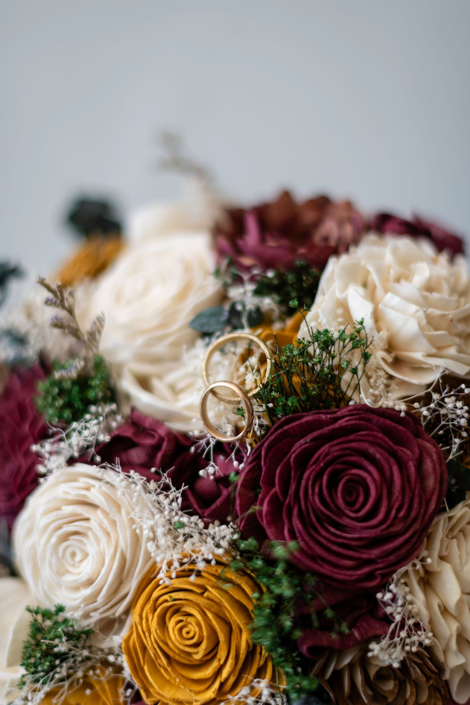 Elegant wedding bouquet with burgundy, cream, and gold roses adorned with two gold wedding bands nestled among the blooms.
