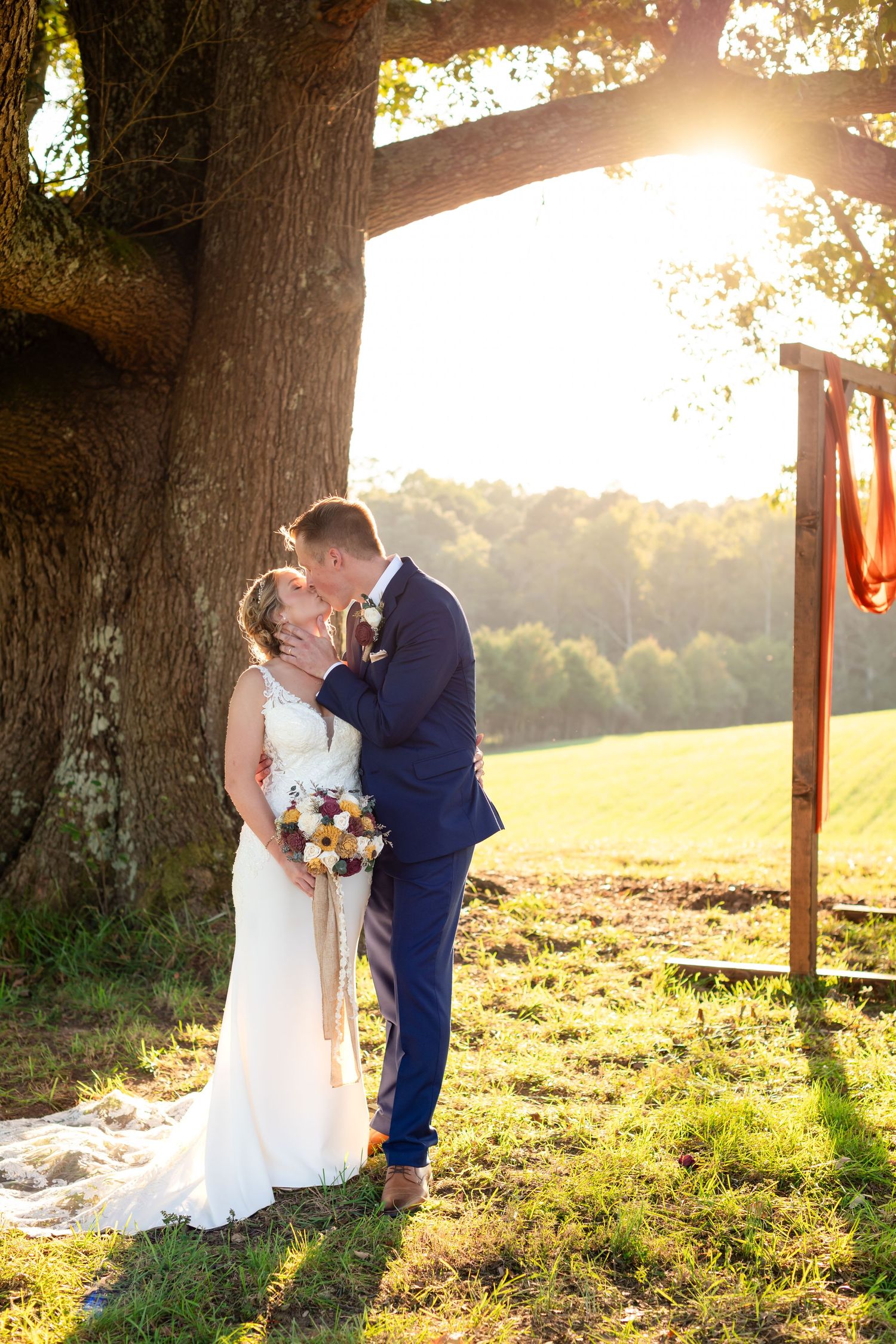 Bride and groom share a sunset kiss under a large oak tree with golden light streaming through the branches.