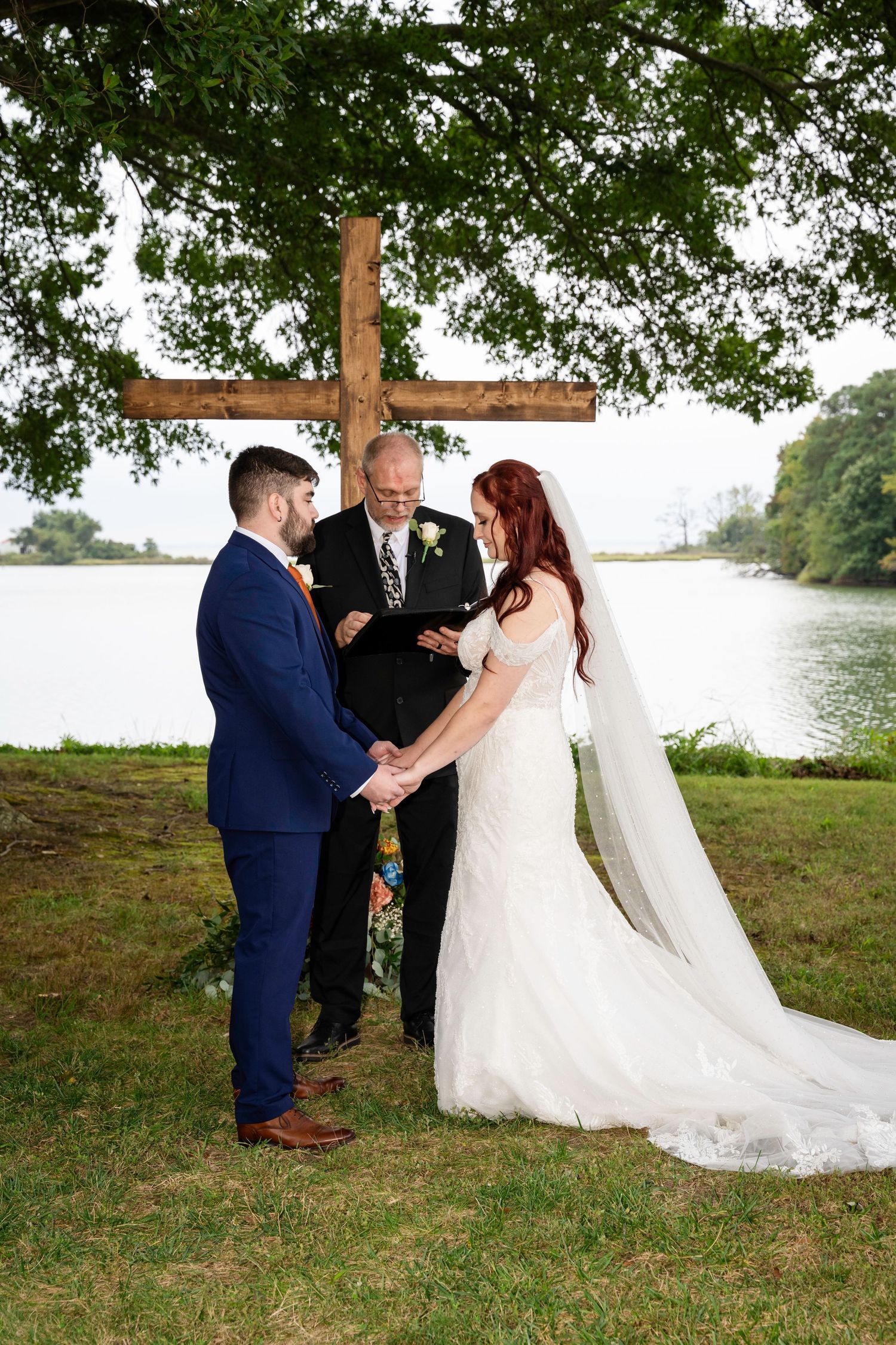 A bride and groom hold hands during their vows before an officiant under a wooden cross by a lake.